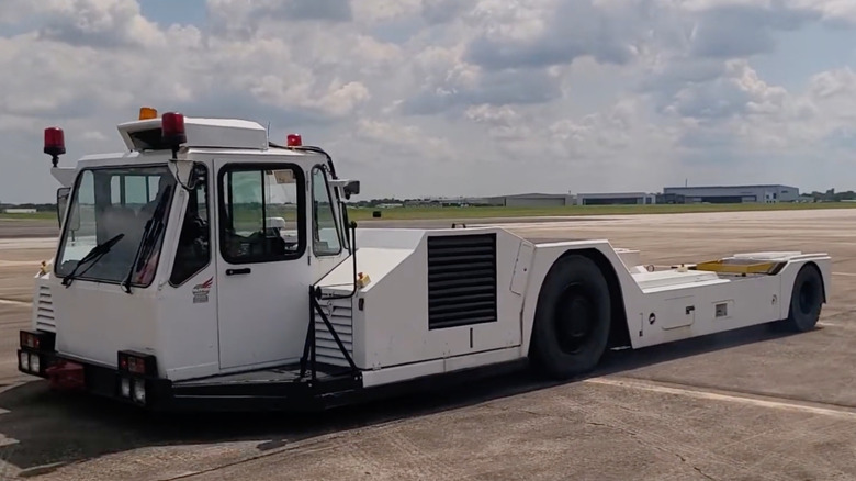 A screenshot showing a front three-quarters shot of a white Tugmaster parked on an airfield in front of a cloudy sky and grass